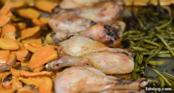 Close-up of seasoned chicken drumsticks and chopped sweet potatoes on a sheet pan, waiting to be roasted