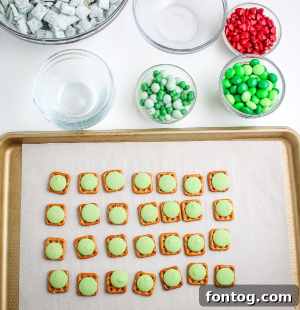 Green candy melts placed on square pretzels on a parchment-lined baking sheet, ready for the oven to create Grinch Pretzel Bites.