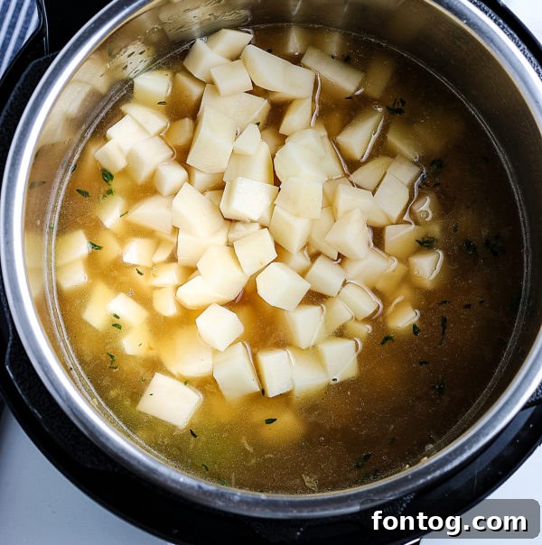 Diced russet potatoes waiting to be added to a pressure cooker pot, surrounded by fresh herbs and other soup ingredients.